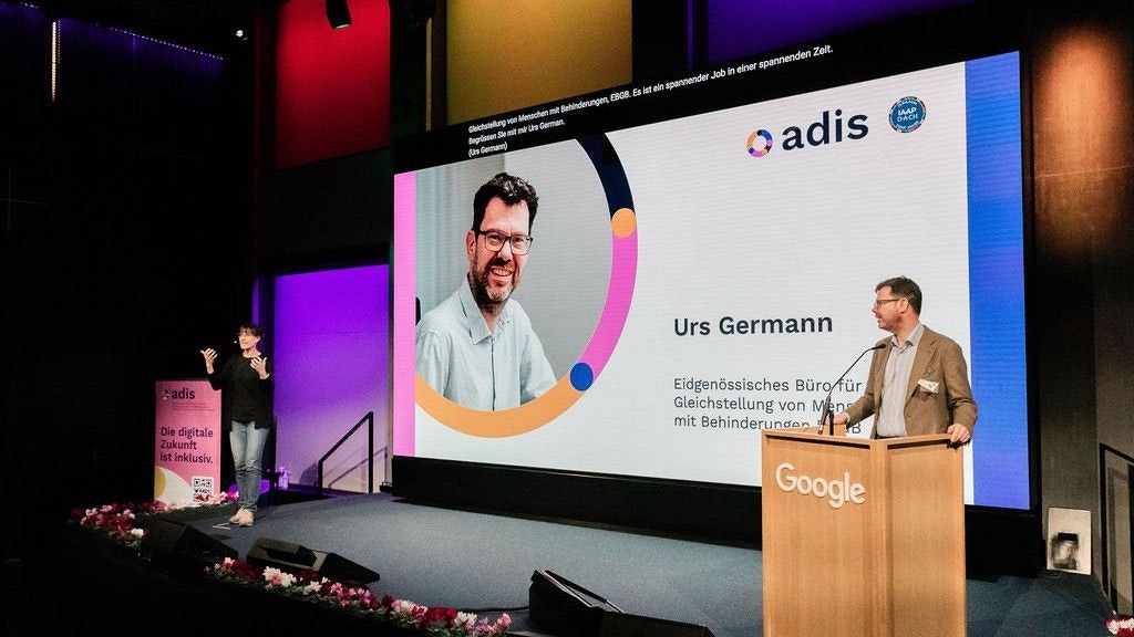 Urs Germann am Rednerpult. Er spricht ins Mikrofon. Im Hintergrund ist sein Bild eingeblendet. Links steht die Gebärdensprachdolmetscherin.