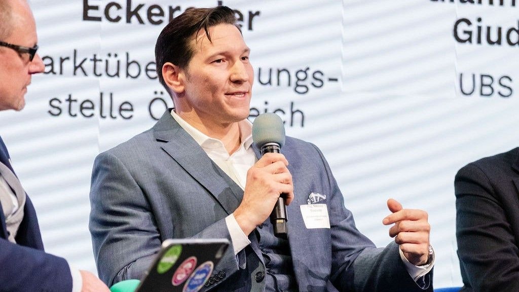 Dr. Stefan Breuer spricht in ein Handmikrofon. Man sieht in sitzend - gross eingeblendet.