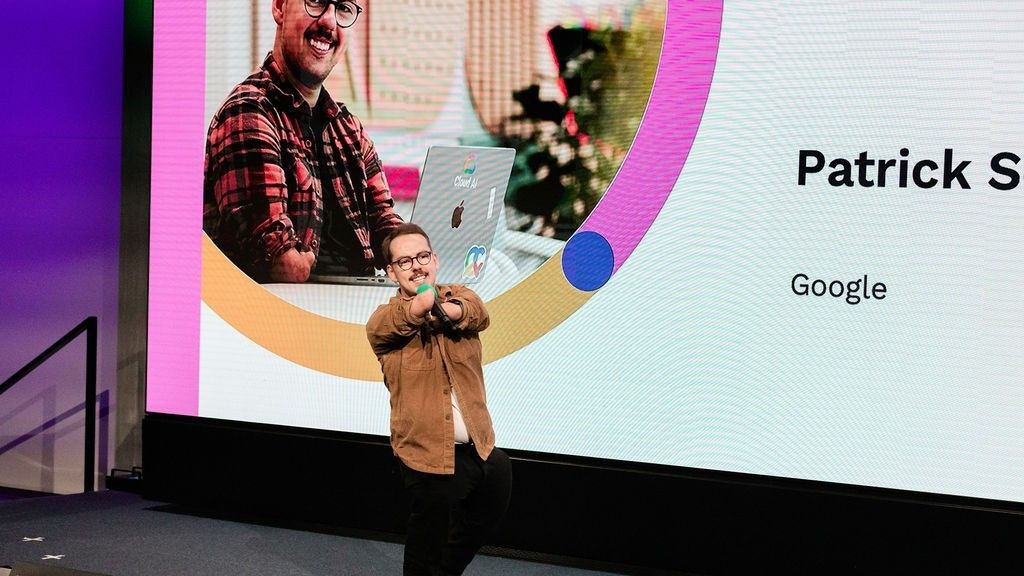 Patrick Schilling auf der Bühne. Er spricht in ein Handmikrofon - im Hintergrund ist sein Bild eingeblendet.