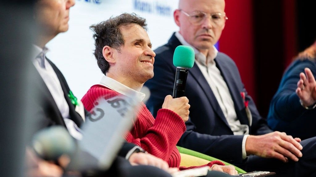 Gianfranco Giudice hält ein Mikrofon in der Hand. Er lächelt. Man sieht ihn sitzend - er ist im Fokus des Bildes. Neben ihm sitzen andere.