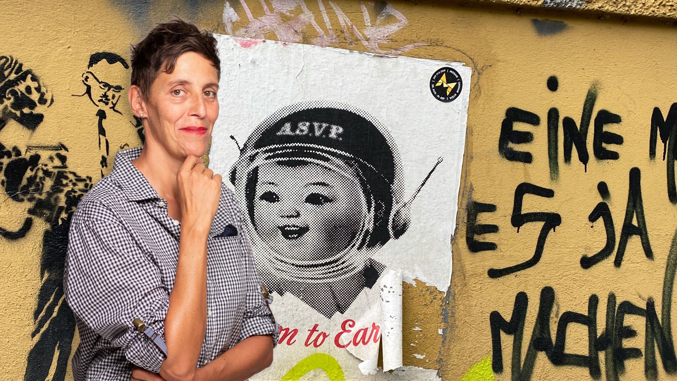 Photo de Sibylle Rau La photo est composée comme un collage artistique. À gauche, on voit Sybille Rau. Elle se tient debout en souriant, la main droite posée sous son menton. Elle a l’air pensive. Le fond est un mur jaune avec une affiche déchirée et plusieurs graffitis. Un des graffitis représente un homme avec une cravate. On peut aussi lire des mots en allemand: «eine», «es» ou «ja». L’affiche représente une fillette portant un casque sur fond blanc.  
