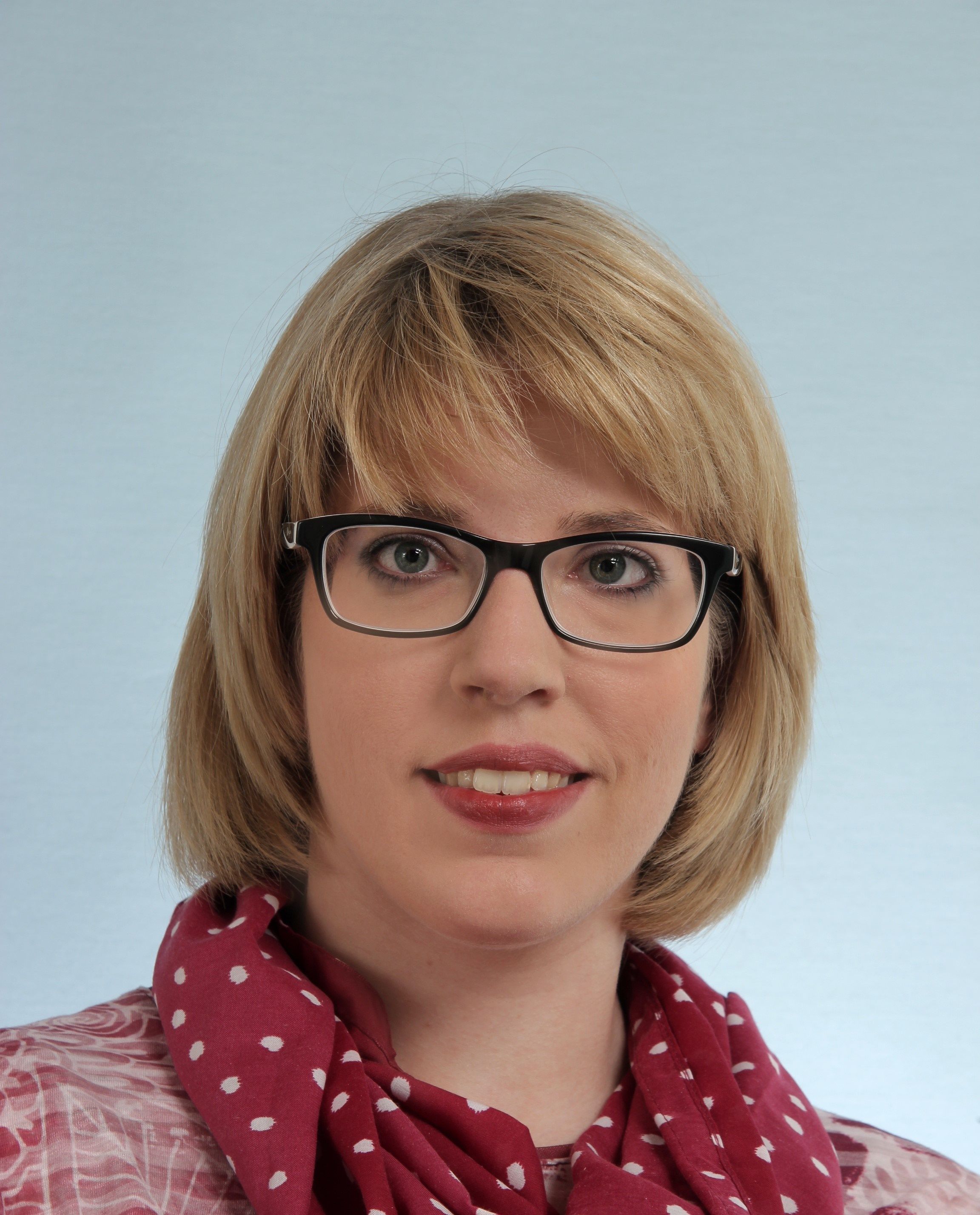 Portrait de Vanessa Leuthold. Elle sourit à l’appareil photo. Madame Leuthold porte des lunettes noires chapeautées par une frange blonde. Elle porte une écharpe rouge à points blancs autour du cou ainsi qu’un haut rouge avec un motif blanc.