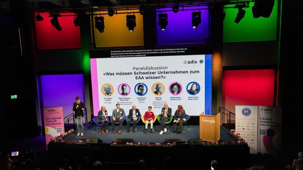 Photo de tous les participants à la table ronde affichée – ils sont tous assis en rangée et ne sont pas reconnaissables de loin. À gauche, l'interprète en langue des signes. À l'arrière-plan, la diapositive avec leurs photos et les carrés de différentes couleurs sur le mur de la pièce.