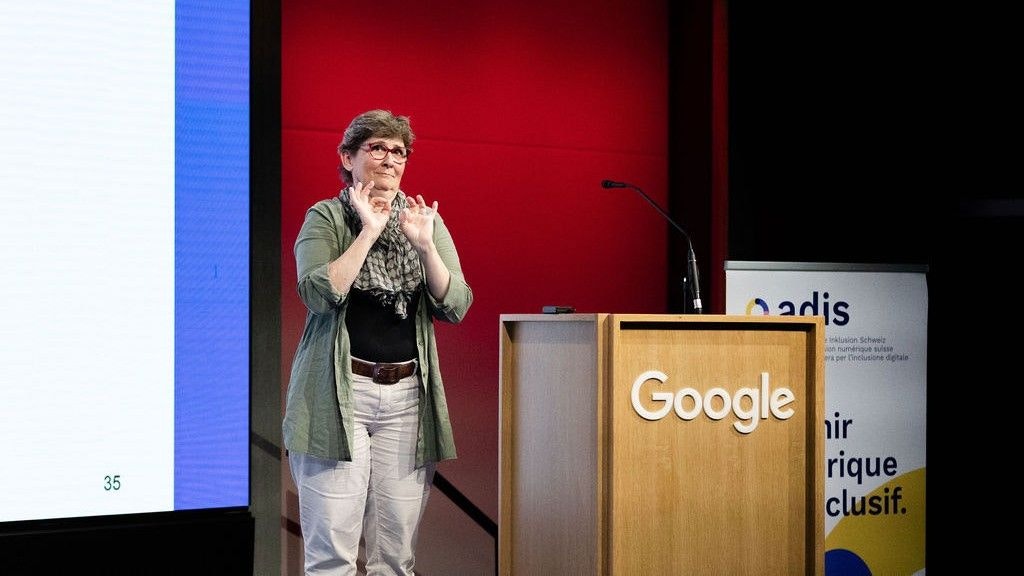 Katja Tissi è in piedi a sinistra del podio. Si vedono le sue mani: sta tenendo la conferenza nella lingua dei segni.