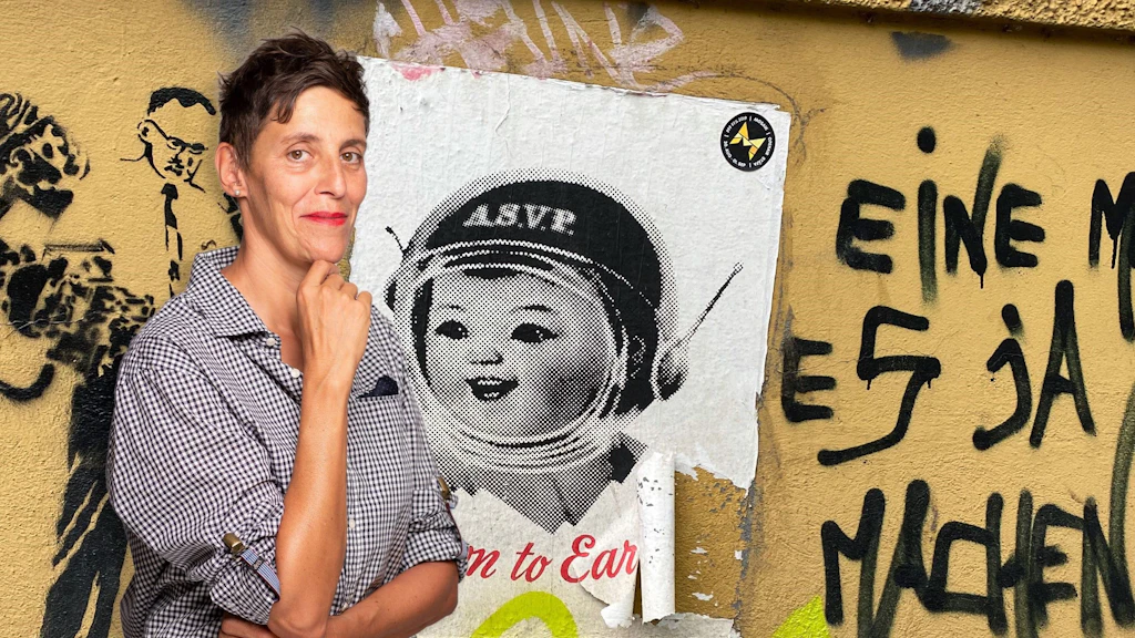Foto di Sibylle Rau La foto è composta come un collage artistico. A sinistra si vede Sybille Rau. È in piedi e sorride, con la mano destra appoggiata sotto il mento. Sembra pensierosa. Lo sfondo è un muro giallo con un manifesto strappato e diversi graffiti. Uno dei graffiti raffigura un uomo con una cravatta. Si possono anche leggere alcune parole in tedesco: «eine», «es» o «ja». Il manifesto raffigura una bambina con un casco su uno sfondo bianco.  