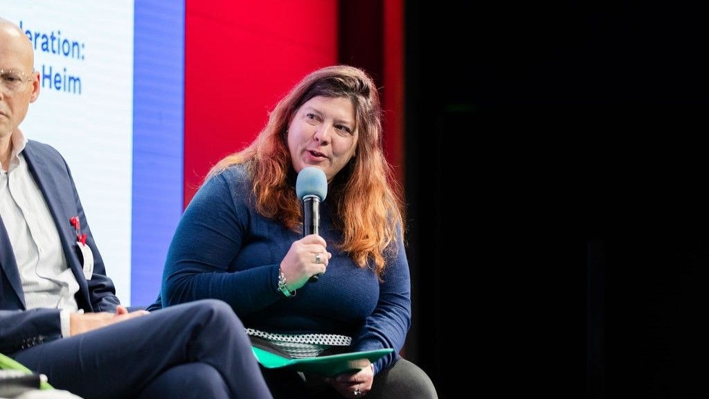 Manu Heim parla al microfono. Il suo sguardo è rivolto a sinistra. L'attenzione è focalizzata su di lei. Accanto a lei ci sono altre persone.