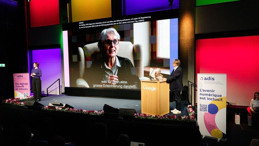 Petra Ritter è in piedi al podio. Alla sua destra c'è un assistente, a sinistra nell'immagine l'interprete della lingua dei segni. Sullo sfondo si vede il suo volto in primo piano.