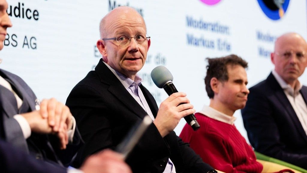 Il dott. Nikolaus Eckereder parla al microfono. Guarda verso sinistra. Lo si vede seduto, l'attenzione è focalizzata su di lui - accanto a lui sono seduti altri.