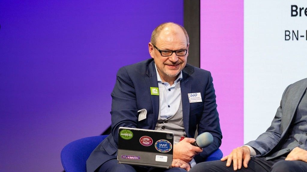 Markus Erle è seduto sulla sedia con il microfono in mano. Sorride. Ha un tablet sulle ginocchia.